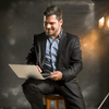 A smiling man holding his laptop sitting on a wood chair